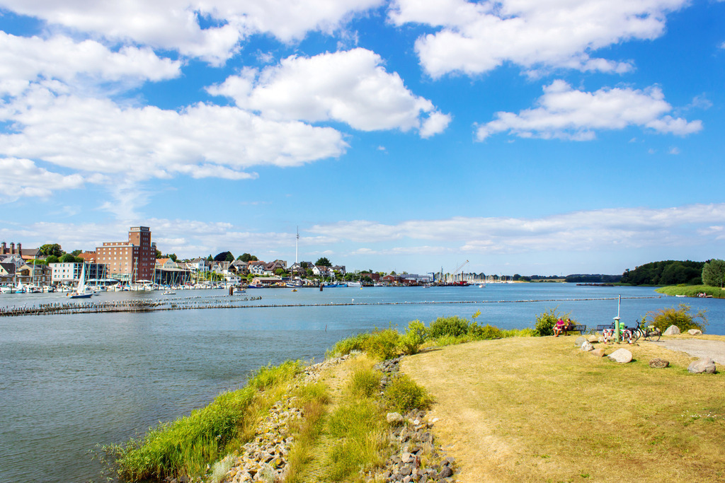 Wandbild: Kappeln an der Schlei im Sommer | Dieses Wandbild im Querformat zeigt die Schlei in Kappeln im Sommer. Im Vordergrund befindet sich das Schleiufer auf der Schwansener Seite der Schlei. Am blauen Himmel befinden sich einige sommerliche Wolken.  - Realisiert mit Pictrs.com
