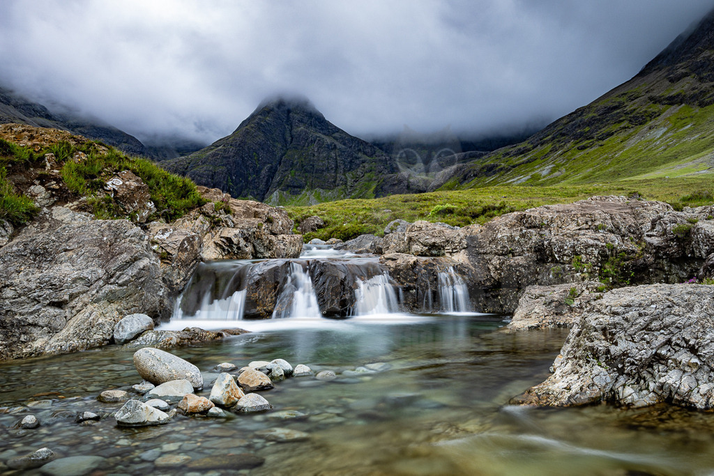 20220721123756 | Diese malerische Aufnahme zeigt die berühmten Fairy Pools auf der Isle of Skye in Schottland. Im Vordergrund fließt ein kristallklarer Fluss über mehrere felsige Stufen und bildet sanfte Kaskaden, deren Wasser durch Langzeitbelichtung seidig weich erscheint. Zahlreiche runde Steine liegen im flachen, durchsichtigen Wasser. Der Flusslauf ist von moosbewachsenen Felsen und üppig grünen Ufern gesäumt. Im Hintergrund erheben sich majestätische, dunkle Berge der Cuillin Range, deren Gipfel teilweise in dichte, mystische Wolken gehüllt sind. Die Berghänge sind mit saftigem Grün bewachsen, was auf die feuchte und kühle Umgebung hindeutet. Die Szene strahlt eine unberührte Wildheit und eine ruhige, fast magische Atmosphäre aus, typisch für die schottischen Highlands an einem bewölkten Sommertag. - Realisiert mit Pictrs.com