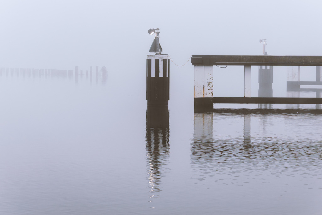 Wandbild: Nebel an der Schleibrücke in Kappeln | Dieses Wandbild im Querformat zeigt das Leuchtfeuer direkt an der Schleibrücke einen nebligen Tag. Auf der Schlei ist eine schöne Spiegelung zu sehen. In der Ferne auf der linken Seite kann man im Nebel den Heringszaun erahnen.  - Realisiert mit Pictrs.com