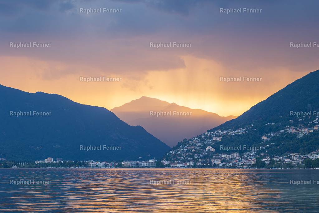 Abendstimmung nach dem Gewitter | Nach einem Sommergewitter über dem Lago Maggiore färbte sich der Himmel in dramatische Orange- und Blautöne. Die Sonnenstrahlen brechen durch die Wolkendecke und tauchen die Berglandschaft in ein faszinierendes Lichtspiel.