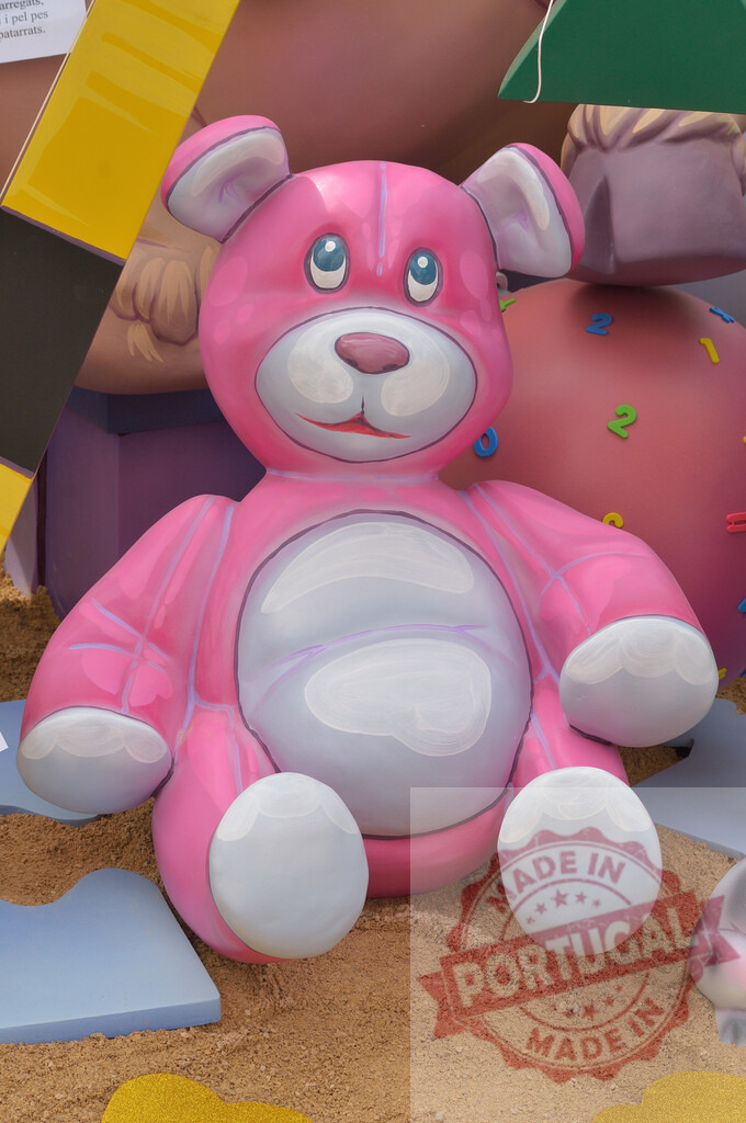 Pink teddy bear sculpture representing childhood memories. Fallas Fiestas Communidad Valenciana | Pink teddy bear sculpture sitting in sand, symbolizing childhood and playfulness - Realized with Pictrs.com
