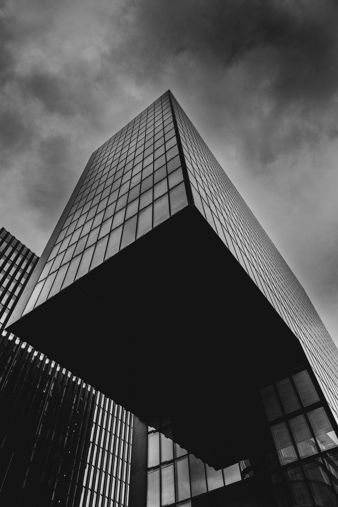 Edge of Gravity – Hyatt Regency Düsseldorf | Eine markante Architekturfotografie in Schwarz-Weiß, aufgenommen aus einer dramatischen Untersicht. Die klare Linienführung, die geometrische Rasterfassade und die massive dunkle Gebäudestruktur erzeugen eine kraftvolle, fast schwebende Wirkung gegen den wolkenreichen Himmel.Eine markante Schwarz-Weiß-Aufnahme des Hyatt Regency im Düsseldorfer MedienHafen, fotografiert aus einer dramatischen Untersicht.Die klare Rasterfassade, scharfen Gebäudekanten und tiefen Schatten erzeugen eine kraftvolle geometrische Komposition. Der dramatische Himmel verstärkt die monumentale Wirkung der modernen Architektur und verwandelt das Gebäude in ein minimalistisches Fine-Art-Motiv voller Spannung, Kontrast und urbaner Präsenz. - Realisiert mit Pictrs.com