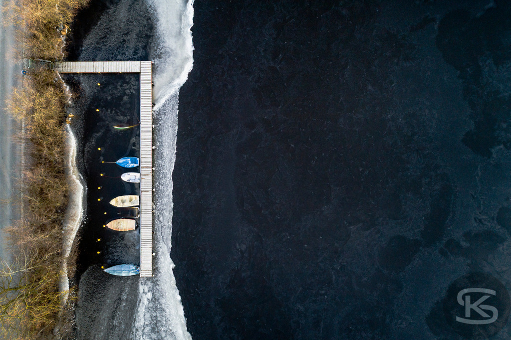 Eingefrorene Boote am Wintersteg – Drohnenaufnahme eines zugefrorenen Sees | Beeindruckende Luftaufnahme von eingefrorenen Booten am Steg eines zugefrorenen Sees. Die Drohnenfotografie zeigt winterliche Stille mit Baumschatten auf der Eisfläche aus der Vogelperspektive – atmosphärische Winterimpression. - Realisiert mit Pictrs.com