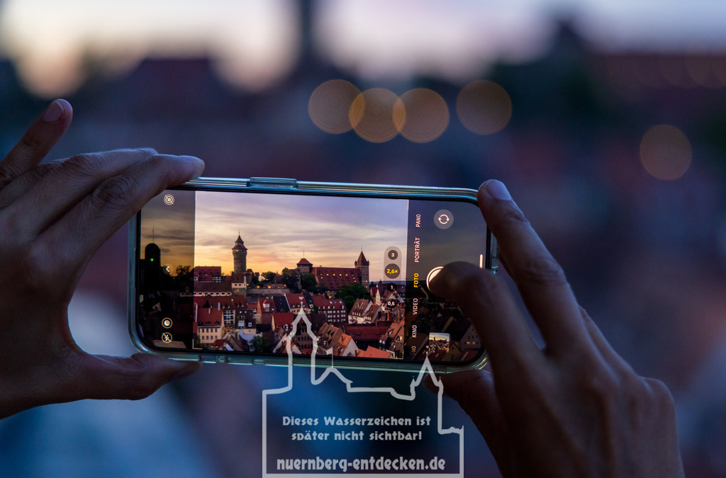 Nürnberger Stadtbild am Abend | Eine Person fotografiert die Nürnberger Altstadt bei Sonnenuntergang mit einem Smartphone. Auf dem Display ist die Kaiserburg in Nürnberg deutlich zu sehen, während der Hintergrund unscharf bleibt und in Bokeh-Lichtern verschwimmt. Die Szene verbindet moderne Technologie mit historischer Architektur. - Realisiert mit Pictrs.com