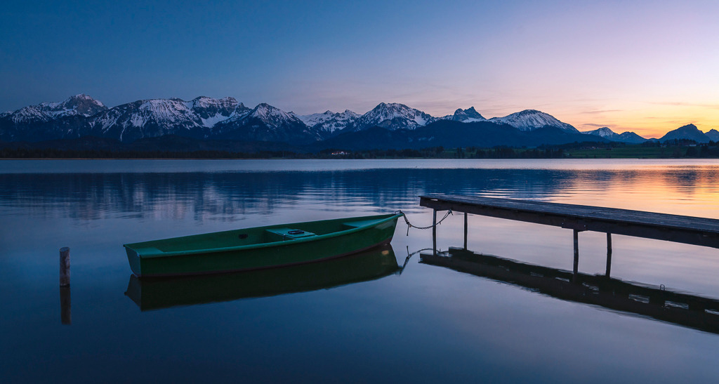 Abendstille – Blick über den Hopfensee | Der Hopfensee im Allgäu liegt ruhig in der blauen Stunde, eingerahmt von den schneebedeckten Alpen. Das Ruderboot und der Steg betonen die friedliche Atmosphäre und laden zum Innehalten ein. - Realisiert mit Pictrs.com