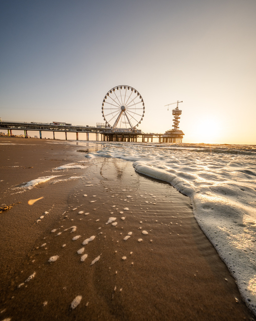 Riesenrad am Strand von Scheveningen | Das Riesenrad auf dem Pier von Scheveningen im warmen Gegenlicht, gesehen aus einer tiefen Perspektive über den weitläufigen Strand. - Realisiert mit Pictrs.com