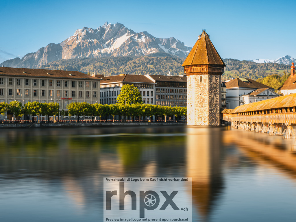 Kapellbrücke und Pilatus im Morgenlicht, Luzern | Die Kapellbrücke mit dem Wasserturm spiegelt sich ruhig in der Reuss, während im Hintergrund der Pilatus im klaren Morgenlicht erscheint. Klassische Luzerner Ansicht mit sauberer Linienführung, warmem Licht und ruhiger Wasseroberfläche – ein zeitloses Postkartenmotiv mit alpinem Bezug. - Realisiert mit Pictrs.com