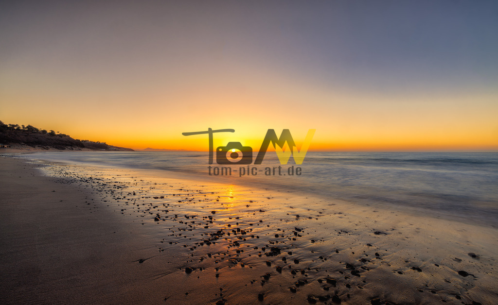 Sonnenaufgang über glitzernde Steine am Strand-Fuerteventura | Ein neuer Tag beginnt , am Strand auf Fuerteventura. Eine Aufnahme mit langer Belichtungszeit. Durch den tiefen Stand der Sonne, leuchten die feuchten Steine orange rot im funkelndem Licht der Sonne. - Realisiert mit Pictrs.com