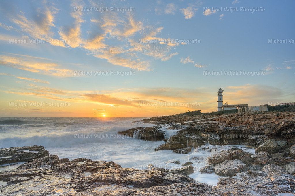 Sonnenuntgang am Cap de ses Salines auf Mallorca | Am Cap de ses Salines zeigt das Foto eine natürliche, unverstellte Küstenszene im warmen Licht des Sonnenuntergangs. Die Felsen im Vordergrund wirken rau und kantig, vom tief stehenden Licht weich betont, während die Brandung mit spürbarer Kraft gegen die Küste schlägt und helle Gischt aufwirbelt. Rechts im Bild steht der Leuchtturm, ruhig und klar sichtbar, als fester Orientierungspunkt inmitten der Bewegung des Meeres. - Realisiert mit Pictrs.com
