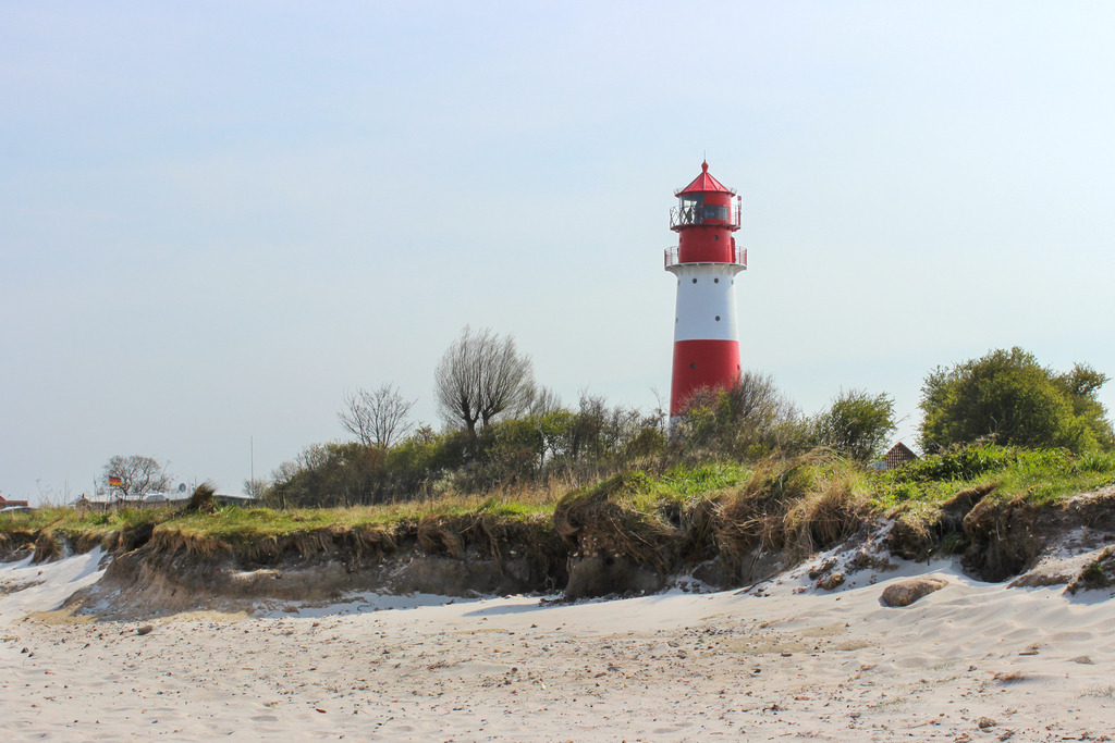 Wandbild: Leuchtturm in Falshöft im Frühling | Dieses Wandbild im Querformat zeigt den Strand und den Leuchtturm in Falshöft im Frühling. Im Vordergrund ist der Strand zu sehen. Der Leuchtturm steht inmitten von Gräsern und Büschen. Der blaue Himmel ist fast wolkenlos.  - Realisiert mit Pictrs.com