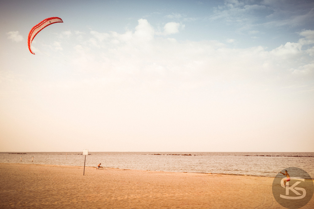 Dynamische Kitesurf-Action an einem sonnigen Strand | Erleben Sie die dynamische Action des Kitesurfens an einem sonnigen Strand am Meer. Ein leuchtend roter Kite schwebt hoch am Himmel über dem weiten Horizont. Am Strand und im Wasser sind Personen zu sehen, die diesen beliebten Wassersport genießen. Das Bild fängt die sommerliche Urlaubsatmosphäre ein und vermittelt ein Gefühl von Leichtigkeit, Freiheit und Windkraft. - Realisiert mit Pictrs.com