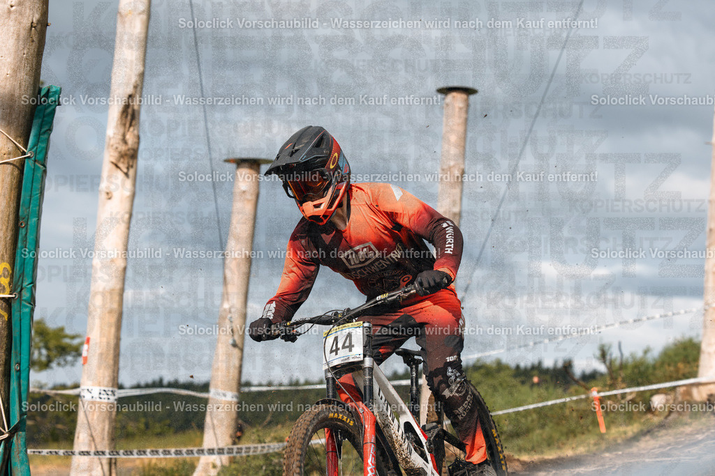 Mountainbike-Action | Ein Mountainbiker in voller Montur rast auf einer unbefestigten Straße entlang. Er trägt einen schwarzen Helm mit roten Akzenten, eine rote Jacke, rote Handschuhe und rote Hose. Auf seinem Rücken befindet sich eine Nummer 44. Das Mountainbike hat eine rote Rahmenfarbe und ist mit schwarzen Reifen und einer schwarzen Federgabel ausgestattet. Die Straße ist staubig und führt durch eine ländliche Umgebung mit Bäumen und Büschen am Straßenrand. Der Himmel ist blau mit wenigen Wolken. - Realisiert mit Pictrs.com