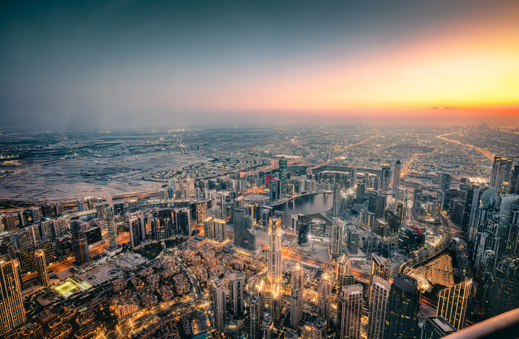 Panoramablick über die Skyline von Dubai bei Sonnenuntergang | Das Bild zeigt einen weiten Panoramablick über die Skyline von Dubai bei Dämmerung oder Sonnenuntergang, aufgenommen aus großer Höhe (vermutlich von einer der Aussichtsplattformen des Burj Khalifa).Der Blick geht über das dichte Netz aus Hochhäusern und Straßenschluchten von Downtown Dubai bis hin zum Horizont und der Wüsten- oder Küstenregion.Am rechten oberen Bildrand zeigt sich der farbenfrohe Übergang eines Sonnenuntergangs, während die Stadt unten bereits mit zahlreichen Lichtern beleuchtet ist. - Realisiert mit Pictrs.com