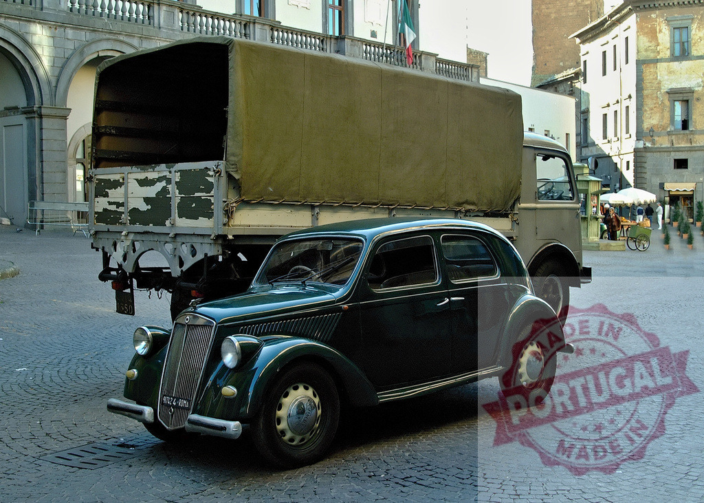Vintage Lancia Ardea car parked in Italian city square | Classic Lancia Ardea from 1940s stands in historic Italian plaza with military truck in background - Realized with Pictrs.com