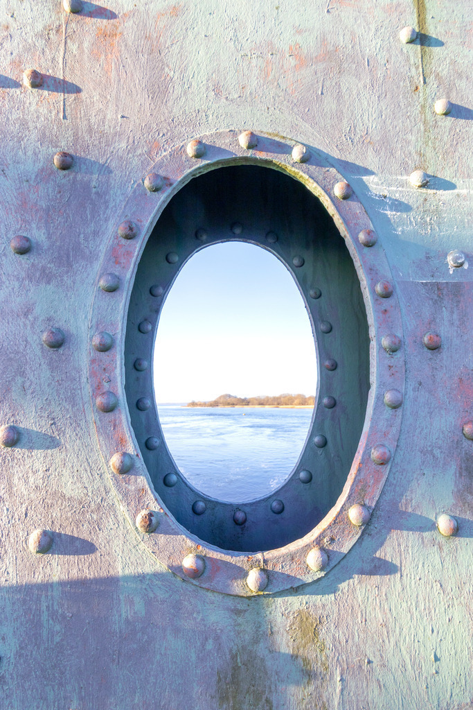 Wandbild: Brückenteil der Lindaunisbrücke als Bilderrahmen | Dieses Wandbild im Querformat zeigt einen Teil der Lindaunisbrücke mit einem Ovalen Loch. Auf dem Brückenteil sind zahlreiche Nieten zu sehen. Durch das Loch im Brückenteil kann man in der Unschärfe die Schlei sowie das Schleiufer sehen. Es wirkt es wäre es ein Bilderrahmen aus Metall.  - Realisiert mit Pictrs.com