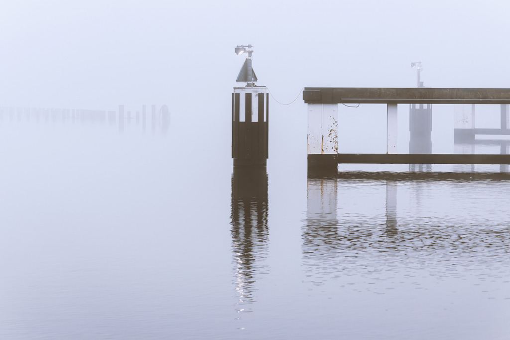Wandbild: Brückeneinfahrt im Nebel – Schleibrücke Kappeln | Ein stilles Wassermotiv an der Schleibrücke in Kappeln: Die Brückeneinfahrt erscheint im dichten Nebel als reduzierte Struktur aus Metall und Holz, deren Linien sich sanft im Wasser spiegeln. Pfähle und Geländer verlieren sich im Dunst, wodurch Wasser und Himmel zu einer weichen, nahezu grenzenlosen Fläche verschmelzen. Die gedämpfte Farbwelt und die minimalistische Komposition verleihen diesem Bild eine ruhige, elegante Fine‑Art‑Ästhetik mit klar erkennbarem regionalem Charakter.