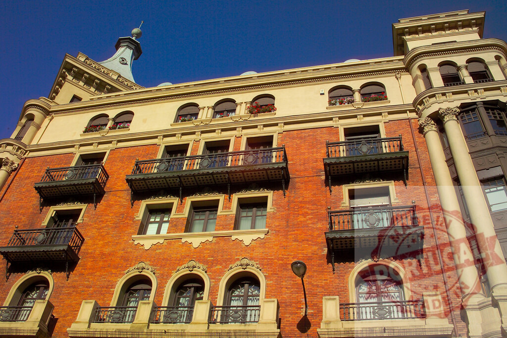 Ornate brick building architecture with balconies and spire in city. Spain - Burgos | Old brick building displaying intricate architectural details, balconies, and a decorative spire against a clear sky - Realized with Pictrs.com