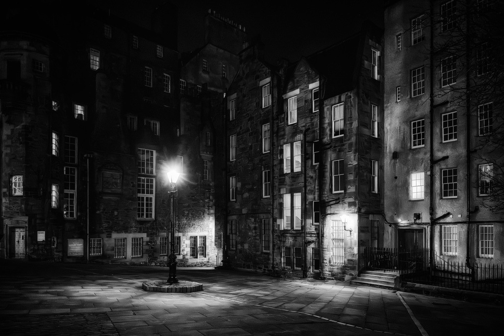 Naechtliche Altstadtgasse, Edinburgh | Eine historische Gasse in Edinburghs Altstadt wird von einer einzelnen Strassenlaterne beleuchtet, deren Licht die Pflastersteine und die Fassaden der alten Gebaeude akzentuiert. Die schwarz-weiss-Darstellung verstaerkt die dramatische Stimmung und hebt die architektonischen Details der Steinbauten hervor, waehrend einige Fenster von innen leuchten. Die Komposition mit der zentralen Laterne und den umgebenden Gebaeuden schafft eine zeitlose, atmosphaerische Szene, ideal fuer Reise- und Kulturthemen. - Realisiert mit Pictrs.com