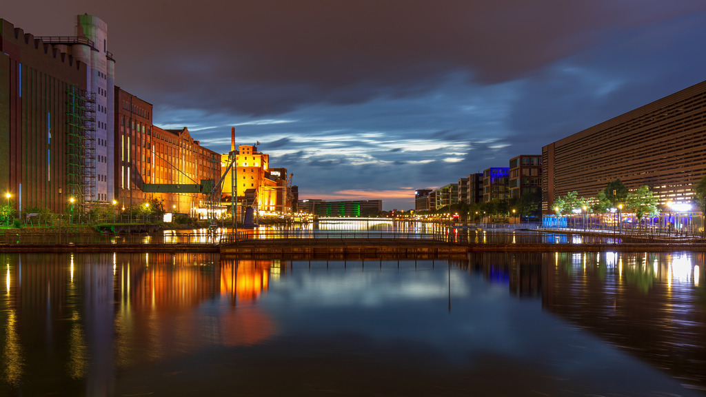 Wandbild - Stadtlichter im Einklang | Dieses faszinierende Bild zeigt eine nächtliche Stadtlandschaft an einem Fluss. Die Gebäude entlang des Ufers sind erleuchtet, was ihren architektonischen Charakter hervorhebt. Links sind historische Industriegebäude zu sehen, während rechts moderne Büro- und Wohngebäude dominieren. Die Spiegelung der bunten Lichter im ruhigen Wasser des Flusses schafft ein lebendiges, doch gleichzeitig beruhigendes Ambiente. Der Himmel ist bewölkt, aber das Licht der untergehenden Sonne erzeugt eine dramatische und malerische Atmosphäre.