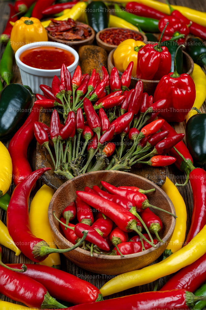 Bunte Chili und Paprika Vielfalt in rustikaler Küche | Diese appetitliche Aufnahme zeigt eine beeindruckende Sammlung verschiedener Chilisorten und bunter Paprika auf rustikalem Holzuntergrund. Frische rote Chilischoten liegen gebündelt neben gelben und grünen Paprikaschoten. Eine weiße Schale enthält rote Chilisauce, während hölzerne Schälchen verschiedene getrocknete Gewürze präsentieren. Die Komposition strahlt Wärme und Authentizität aus. Perfekt für Kochbücher, Gewürzhandel oder kulinarische Magazine. Die natürliche Beleuchtung betont die leuchtenden Farben und die unterschiedlichen Texturen der Gewürze. - Realisiert mit Pictrs.com