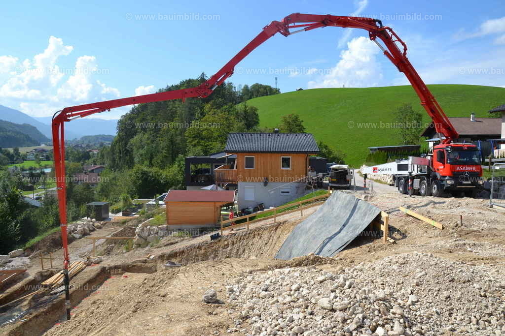 Betonpumpe | Betonpumpe mit roten Auslegern oberhalb Baugrube in Hanglage, Hintergrund grüne Berglandschaft, blauer Himmel, Wolken  