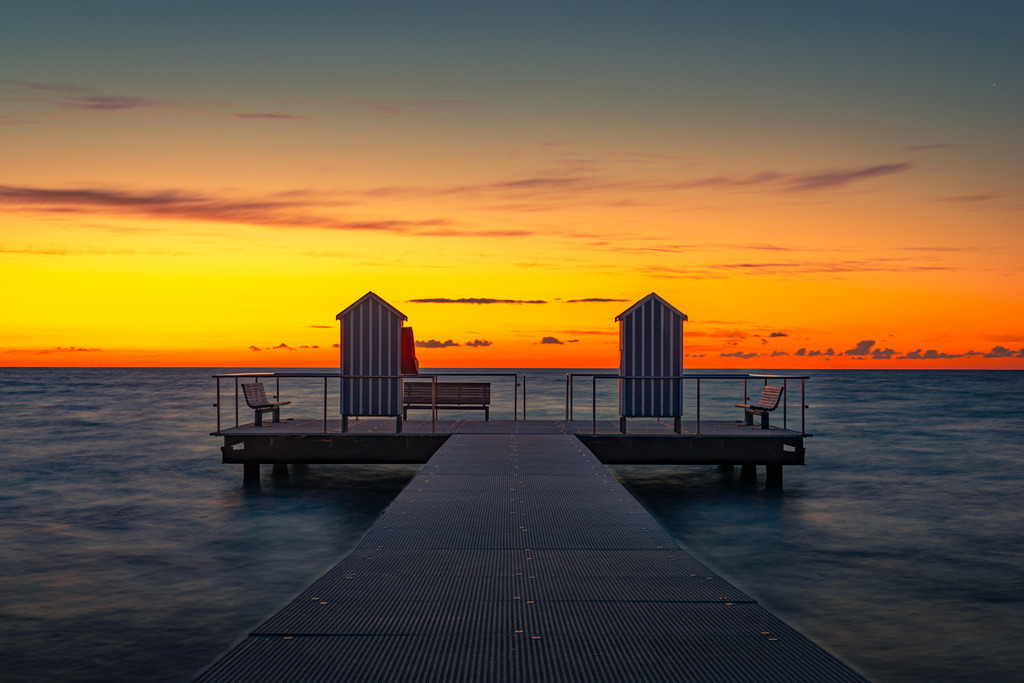 Badesteg | Der Badesteg in der Ostsee im Örtchen Stein bei Laboe erstreckt sich weit ins ruhige Wasser und lädt zum Verweilen und Genießen eines friedlichen Sonnenuntergangs ein. Das orange-goldene Licht der untergehenden Sonne taucht die Umgebung in ein warmes Licht. Eine friedliche Atmosphäre erfüllt die Szenerie. Ein Ort der Ruhe und Entspannung, um den Tag hinter sich zu lassen und in die Nacht zu gleiten.