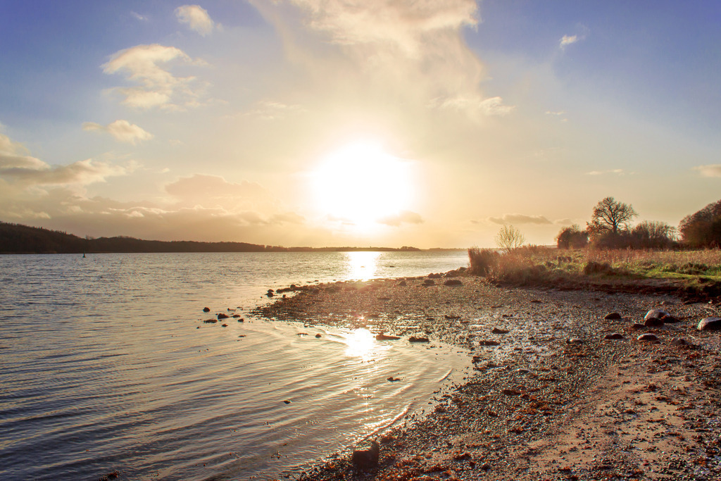 Wandbild: Sonnenuntergang am Ostseefjord | Dieses Wandbild im Querformat zeigt einen schönen Sonnenuntergang am Ostseefjord. Die tiefstehende Sonne spiegelt sich auf der Schlei. Im Vordergrund ist der Schleistrand zu sehen. Am blauen Himmel gibt befinden sich einige Wolken. - Realisiert mit Pictrs.com