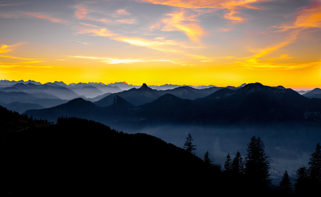 Silhouette der bayrischen Alpen während eines idyllischen Sonnenuntergangs am Tegernsee | Das Bild zeigt einen Sonnenuntergang über einem Gebirge, wobei es sich bei dem markanten Gipfel in der Mitte .Die Umgebung bietet vielfältige Möglichkeiten für Wanderungen und Erholung in den Bergen. Die Szene zeigt eine typische Schichtstruktur von Bergrücken im Dunst.  - Realisiert mit Pictrs.com