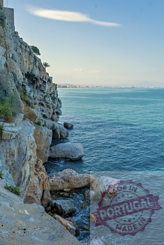 Rocky coast and Mediterranean sea with distant city | Rocky cliffside meeting the calm sea with a distant urban skyline under a clear sky - Realized with Pictrs.com