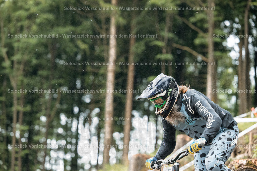 Mountainbike-Abenteuer im Regen | Ein Mountainbiker in voller Ausrüstung rast einen steilen, bewaldeten Hang hinab. Er trägt einen Helm mit einer Visier, eine Schutzbrille, einen schwarzen Pullover mit dem Schriftzug 'Anglerstube' und eine Hose mit einem auffälligen Muster. Er greift die Lenker eines Mountainbikes und scheint sich auf die Fahrt zu konzentrieren. Der Hintergrund besteht aus dichtem Grün und Bäumen, was auf eine natürliche Umgebung hindeutet. Es regnet leicht, was die Atmosphäre feucht und dynamisch macht. - Realisiert mit Pictrs.com