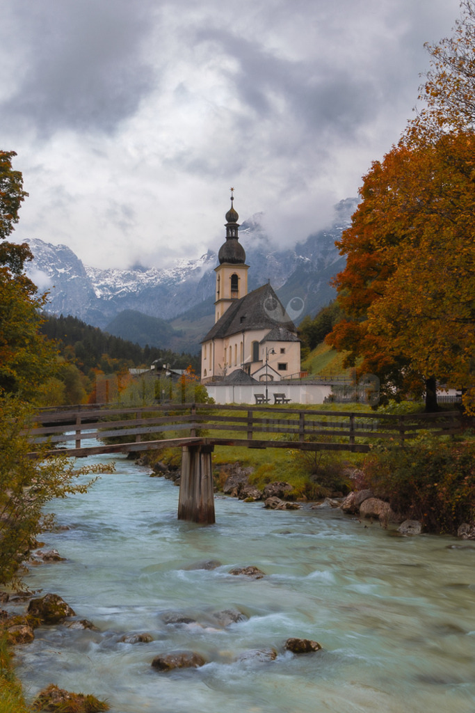 R5M20129_20251009 | Ein malerisches Hochformat der berühmten Pfarrkirche St. Sebastian in Ramsau bei Berchtesgaden, Bayern. Im Vordergrund fließt die Ramsauer Ache unter einer rustikalen Holzbrücke hindurch. Die Kirche mit ihrem markanten Zwiebelturm steht auf einer Anhöhe, umgeben von einer niedrigen Mauer und bunten Herbstbäumen. Im Hintergrund erheben sich majestätische, teilweise schneebedeckte Berge unter einem bewölkten Himmel, was der Szene eine dramatische und zugleich friedliche Atmosphäre verleiht. Die Herbstfarben der Bäume sind leuchtend orange und gelb. - Realisiert mit Pictrs.com
