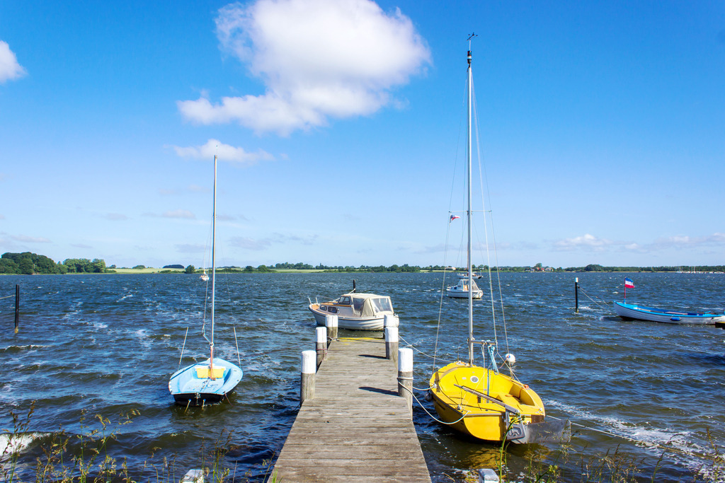 Wandbild: Steg in Maasholm an der Schlei | Dieses Wandbild im Querformat zeigt einen schönen Bootssteg in Maasholm an der Schlei. Direkt am Steg befinden sich zwei Segelboote. Am blauen Himmel sind nur wenige Wolken zu sehen.  - Realisiert mit Pictrs.com