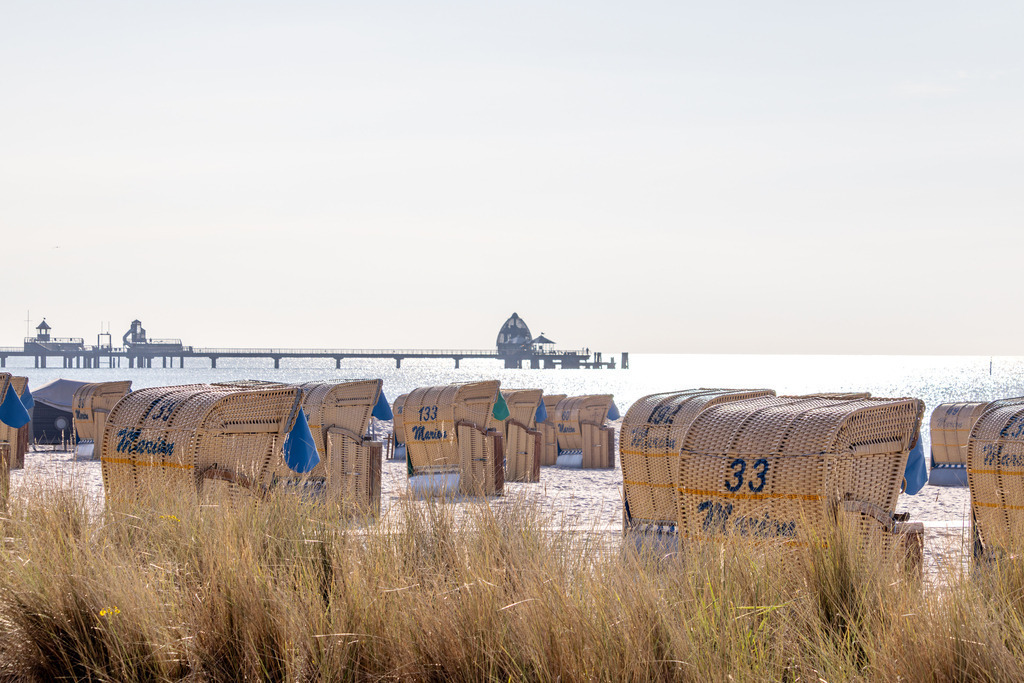 Wandbild: Strandkörbe vor der Seebrücke – Grömitz | Dieses maritime Wandbild zeigt eine ruhige Szene am Strand von Grömitz. Zahlreiche Strandkörbe stehen ordentlich aufgereiht im Sand und prägen die typische Küstenkulisse. Im Hintergrund erstreckt sich die Seebrücke von Grömitz weit ins Meer hinaus, gekrönt von der markanten Tauchglocke – einem Wahrzeichen des Ortes und beliebten Ziel für Besucher. Der Strand wird im Vordergrund von hohem Dünengras eingerahmt, das dem Bild eine natürliche Struktur verleiht. Das Wasser liegt ruhig, und der helle Himmel sorgt für eine freundliche, klare Atmosphäre. Dieses Motiv verbindet Ordnung und Weite mit dem typischen Ostseecharme und eignet sich ideal als Wandbild für maritime Wohnkonzepte. Erhältlich als Leinwandbild, Acrylglasbild, Alu-Dibond FineArt Print oder als Akustikbild – ein stilvoller Blickfang für Zuhause, Büro oder Ferienwohnung. - Realisiert mit Pictrs.com