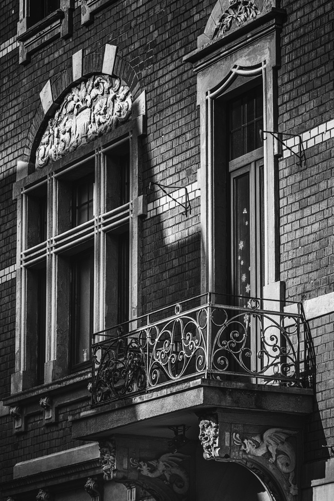Balkon der Stille | Ein architektonisches Detail aus Duisburg, das die Handschrift vergangener Baukunst trägt. Feine Ornamente, geschwungene Metallarbeiten und das Spiel aus Licht und Schatten verleihen dieser historischen Fassade eine zeitlose Eleganz. Die Schwarz-Weiß-Umsetzung lenkt den Blick auf Strukturen, Formen und die stille Schönheit urbaner Architektur. - Realisiert mit Pictrs.com