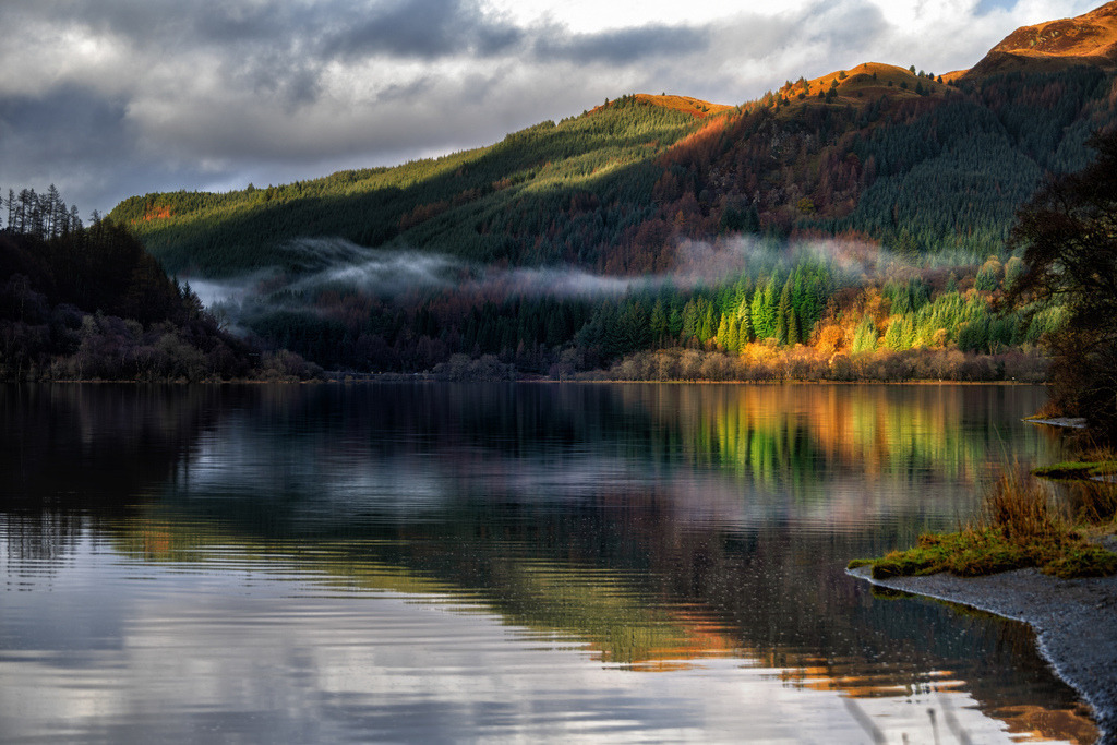 Loch Lubnaig: Herbstliches Lichtspiel | Ein ruhiger See spiegelt die umliegenden, dicht bewaldeten Hügel wider, die teilweise von Sonnenstrahlen durchbrochen werden. Nebelschwaden liegen zwischen den Bäumen und erzeugen eine malerische Tiefenwirkung, während das herbstliche Laub in warmen Farben leuchtet. Die Komposition betont die natürliche Schönheit der schottischen Landschaft mit klaren Reflexionen auf der Wasseroberfläche. - Realisiert mit Pictrs.com