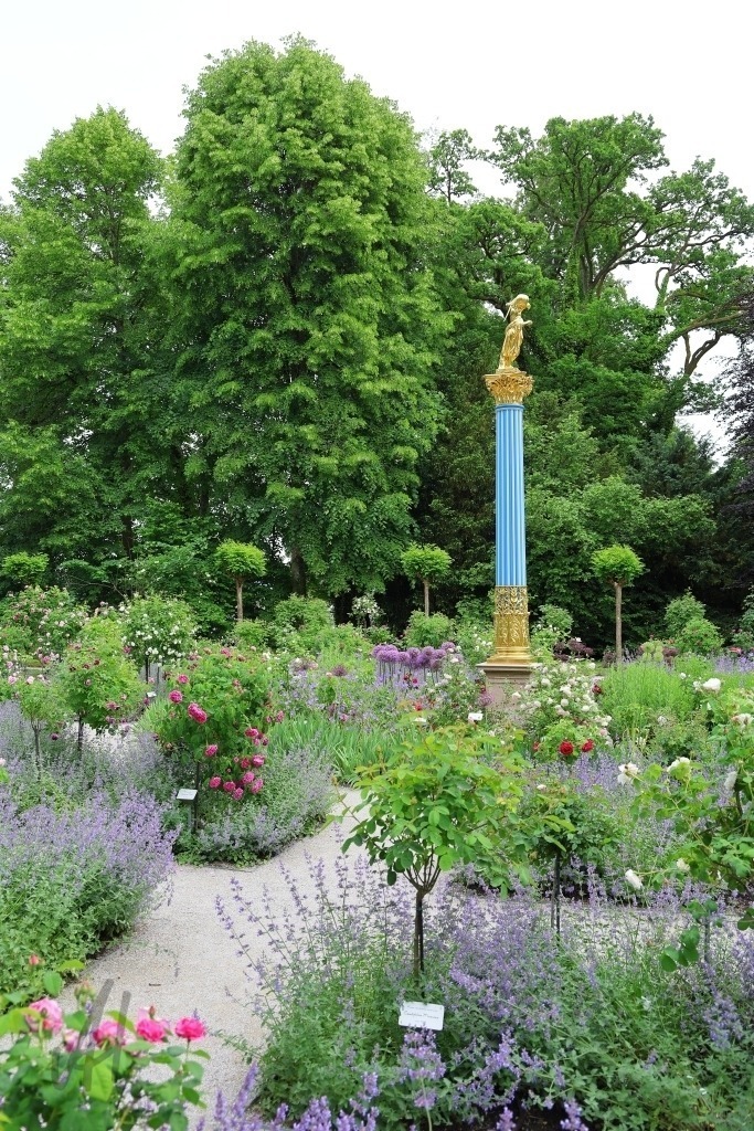 Roseninsel im Starnberger See | Im idyllischen Rosengarten auf der Roseninsel im Starnberger See inmitten sorgfältig angelegter Beete mit blühenden Rosen erhebt sich der goldene Engel als strahlender Blickfang der Anlage. Seine vergoldete Oberfläche reflektiert das Sonnenlicht und setzt einen leuchtenden Akzent zwischen den Farben der Blumen und dem satten Grün der Gartenanlage. Die Stimmung des Bildes wirkt ruhig und zugleich festlich. Der goldene Engel verleiht dem Ort eine besondere Würde und macht den Rosengarten zu einem der schönsten Motive der Roseninsel. - Realisiert mit Pictrs.com