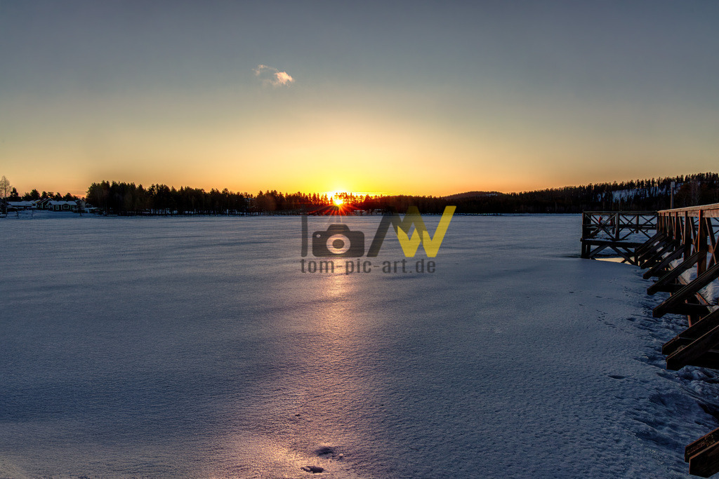 Sonnenuntergang auf zugefrorenen See in Schweden-Kalt--Eis | Das Bild zeigt einen Sonnenuntergang oder Sonnenaufgang über einer zugefrorenen, schneebedeckten Landschaft, die in Schweden liegt. Die Szene strahlt eine ruhige, winterliche Atmosphäre aus und fängt einen Moment natürlicher Schönheit ein.Am rechten Bildrand ist ein hölzerner Steg oder eine Anlegestelle zu sehen, die in den gefrorenen See hineinragt.Im Hintergrund, am - Realisiert mit Pictrs.com