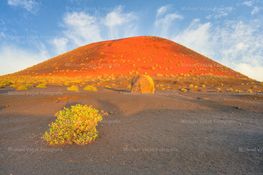 Montana Colorada auf Lanzarote | Die gewaltige Vulkanbombe am Fuß der Montaña Colorada wirkt wie ein stummer Wächter der Landschaft. Dahinter erhebt sich der Vulkan selbst, dessen Flanken im Licht der aufgehenden Sonne in intensiven Rottönen erstrahlen. Ein Bild, das die Kraft der Natur und die stille Schönheit Lanzarotes eindrucksvoll vereint. - Realisiert mit Pictrs.com