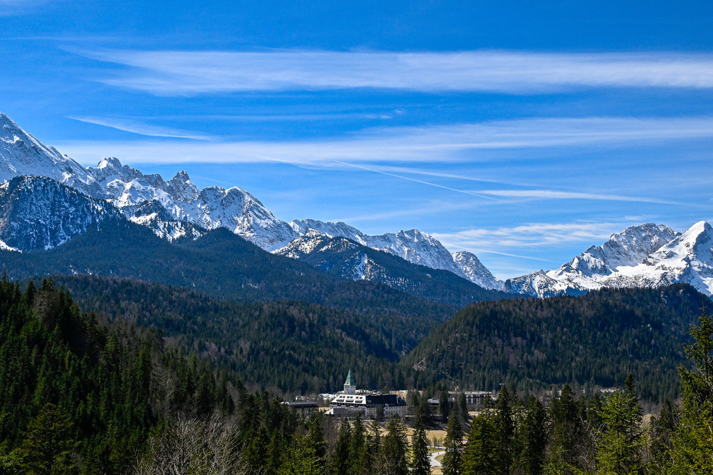 Wo die Berge den Himmel küsse | Ein majestätisches Panorama entfaltet sich über dem Werdenfelser Land: Wie ein stiller Wächter thront ein einsames Schloss mit grünem Turm inmitten eines endlosen Meeres aus dunklen Tannenwäldern. Darüber erhebt sich die atemberaubende Kulisse im Zugspitzgebiet, wo das mächtige Zugspitzmassiv und Deutschlands höchster Berg stolz in den strahlend blauen Himmel ragen. Nahe Garmisch-Partenkirchen gelegen, weckt dieses alpine Idyll eine tiefe Sehnsucht nach Freiheit und erfüllt das Herz mit unendlicher Ruhe. Im Vordergrund Hotel Schloss Elmau. - Realisiert mit Pictrs.com