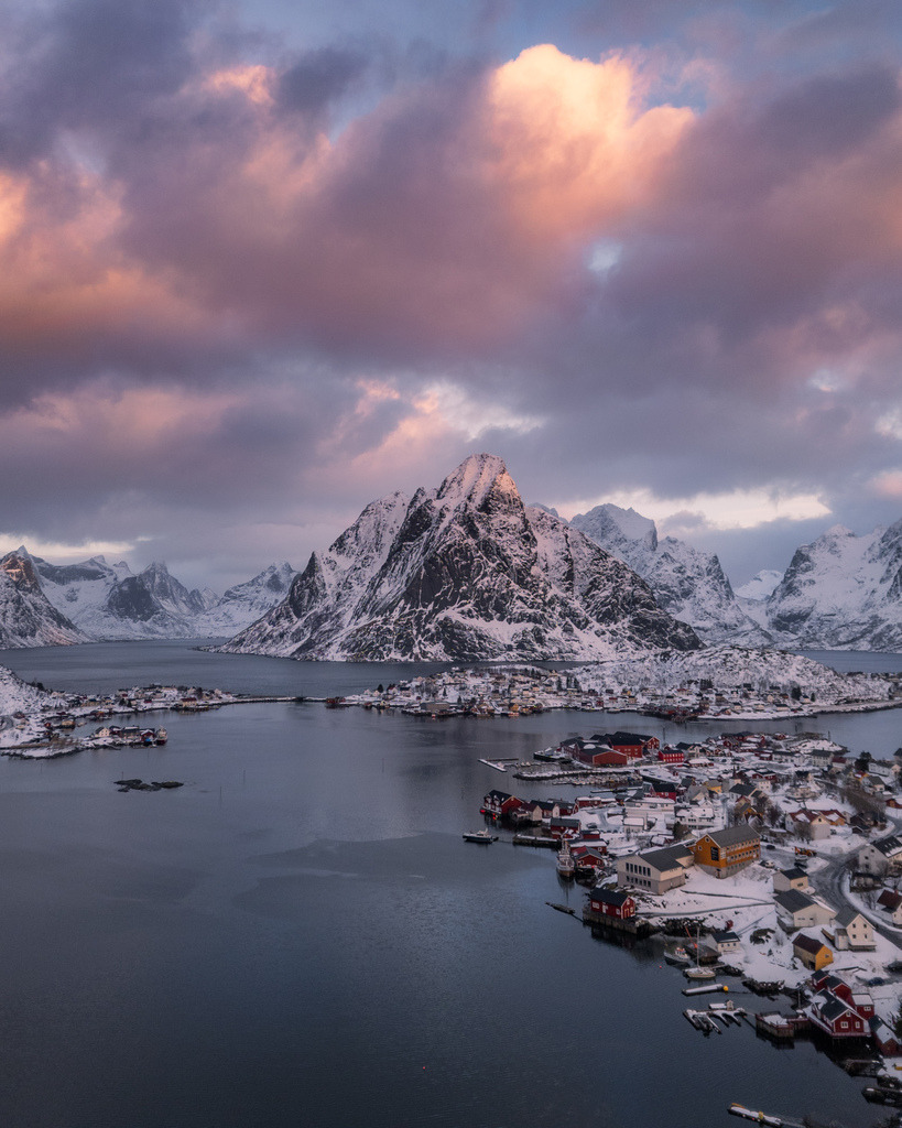 Norwegisches Fischerdorf unter farbigem Winterhimmel | Ein kleiner Küstenort liegt unter einem markanten verschneiten Berg, während der Himmel in warmen Rosa- und Orangetönen leuchtet. Die ruhige Wasserfläche verstärkt die ausgewogene Winterstimmung. - Realisiert mit Pictrs.com