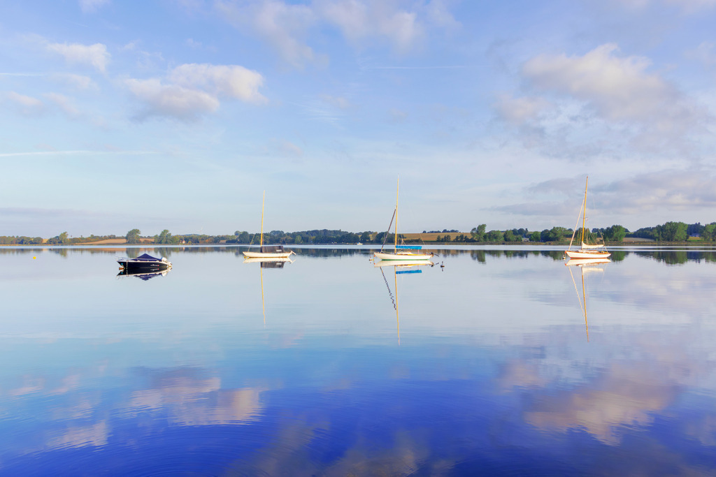 Wandbild: Segelboote spiegeln sich auf der Schlei | Ein friedlicher Morgen an der Schlei – dieses Wandbild fängt die ruhige Atmosphäre der Wasserlandschaft perfekt ein. Die sanften Spiegelungen der Segelboote und der leicht bewölkte Himmel auf der stillen Wasseroberfläche erzeugen eine harmonische und weitläufige Bildkomposition. Die beruhigenden Farben und das sanfte Licht machen dieses Motiv zu einer idealen Ergänzung für jedes Zuhause, das Ruhe und maritime Eleganz ausstrahlen soll. - Realisiert mit Pictrs.com