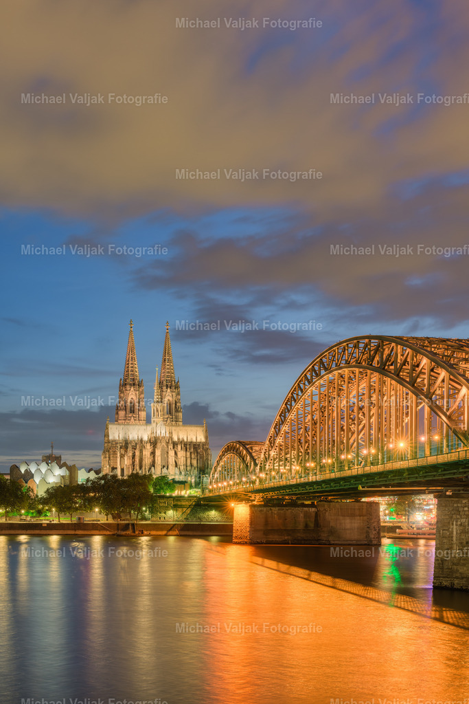 Kölner Dom am Abend | Der Kölner Dom, ein Meisterwerk gotischer Architektur, erstrahlt am Abend in einem beeindruckenden Licht, das seine filigranen Details und die majestätische Struktur hervorhebt. Zusammen mit der Hohenzollernbrücke, die sich mit ihrer robusten Eleganz über den Rhein spannt, bildet er ein ikonisches Panorama, das Besucher aus aller Welt anzieht. Diese beiden Wahrzeichen sind nicht nur bedeutende historische und kulturelle Symbole, sondern auch Zeugen der lebendigen Geschichte und des Fortschritts der Stadt Köln. - Realisiert mit Pictrs.com