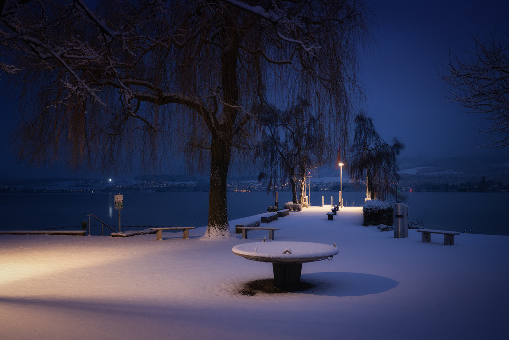 Wintermorgen in Hünibach | Eine schneebedeckte Promenade am Thunersee in Hünibach erstreckt sich unter einem tiefblauen Winterhimmel. Künstliche Beleuchtung taucht den frisch gefallenen Schnee in warmes Licht, während die kahlen Äste einer Trauerweide die Szenerie prägen. Im Hintergrund sind die Lichter der gegenüberliegenden Uferseite sichtbar, die sich im ruhigen Wasser spiegeln. - Realisiert mit Pictrs.com