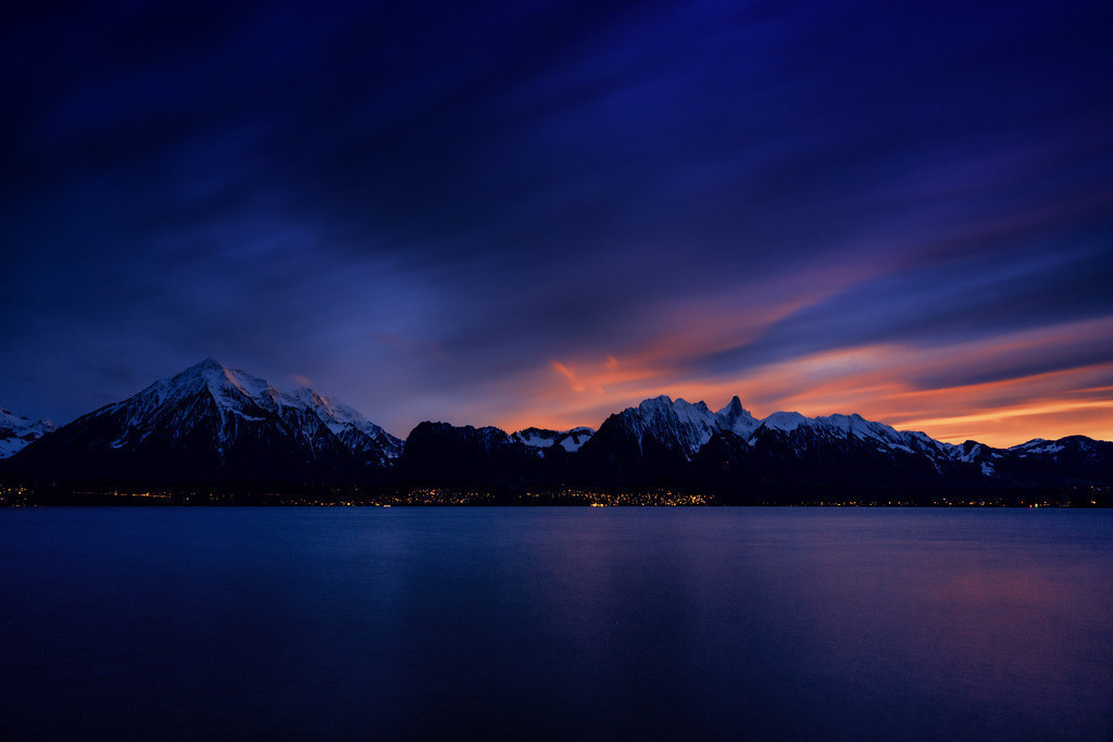 Magie der blauen Stunde | Ein weites Panorama bei Dämmerung zeigt einen ruhigen See im Vordergrund, dessen Oberfläche in tiefem Blau und Violett schimmert. Am Horizont erstreckt sich eine Reihe dunkler, schneebedeckter Berge, darunter der markante Niesen und das Stockhorn, deren Gipfel vom letzten Tageslicht sanft beleuchtet werden. Entlang des Ufers leuchten zahlreiche kleine Lichter von Ortschaften. Der Himmel darüber ist ein dramatisches Farbenspiel aus tiefem Nachtblau, das in der Mitte in leuchtende Orangerot- und Violetttöne eines Sonnenuntergangs übergeht, mit langgezogenen Wolkenstreifen, die sich über den Horizont erstrecken. - Realisiert mit Pictrs.com