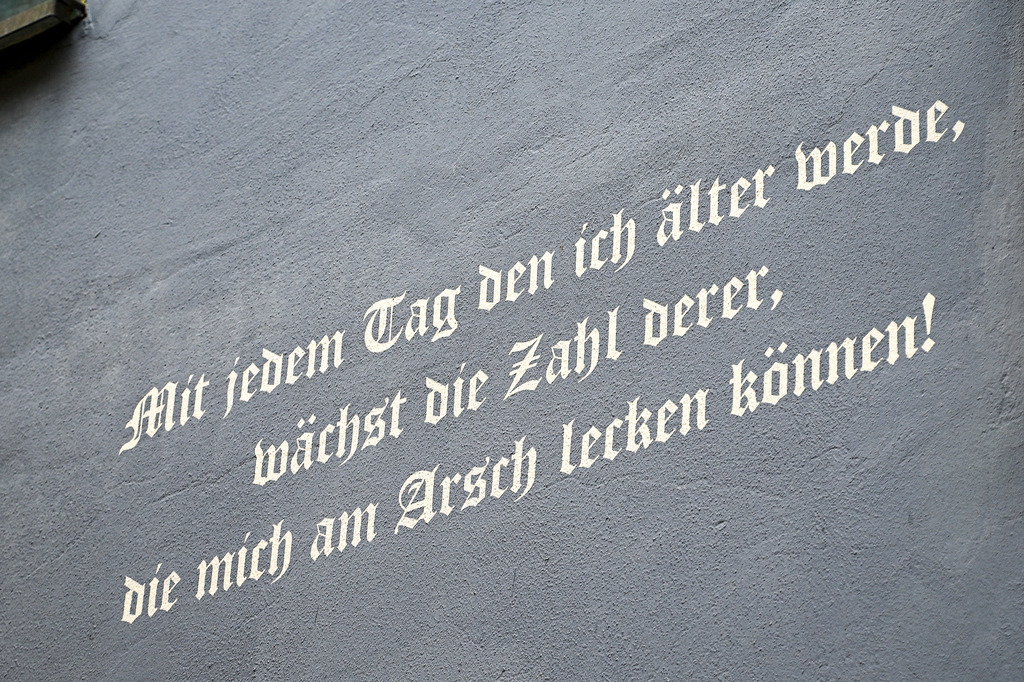 Gelassenheit des Alters: Ein humorvolles Bekenntnis zur Freiheit | Ein Anblick, der zum Schmunzeln anregt und ein befreiendes Gefühl von ehrlicher Nonkonformität vermittelt: In kunstvoller, weißer Frakturschrift prangt ein ironischer Spruch auf einer rauen, graublauen Hauswand. „Mit jedem Tag den ich älter werde, wächst die Zahl derer, die mich am Arsch lecken können!“ steht dort geschrieben. Das Foto fängt die charmante Rebellion gegen gesellschaftliche Zwänge und den humorvollen Gewinn an innerer Freiheit ein, der oft mit den Jahren kommt. Ein augenzwinkerndes Plädoyer für mehr Selbstbestimmung, Gelassenheit und den Mut, die Dinge einfach mal locker zu nehmen. - Realisiert mit Pictrs.com