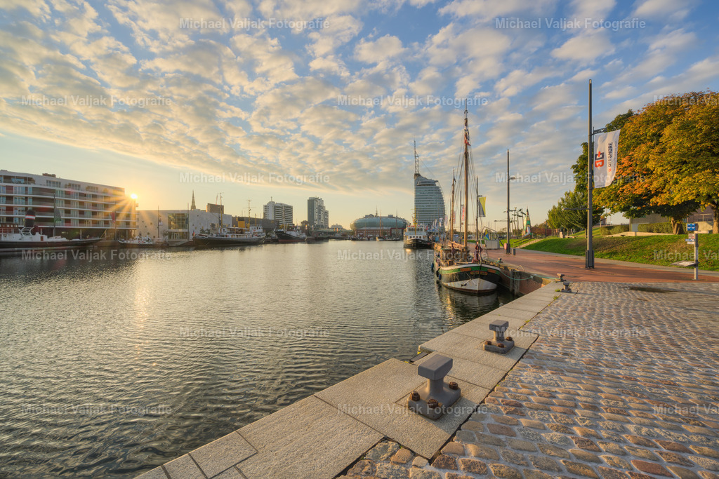 Bremerhaven am Morgen | Vom Kai des Neuen Hafens fällt der Blick auf das Klimahaus und das ATLANTIC Hotel Sail City, deren markante Formen sich gegen den hell werdenden Himmel abzeichnen. Hinter den Gebäuden ist die Sonne gerade aufgegangen – ihr Licht bricht zwischen den Kanten hervor und bildet einen klaren Sonnenstern. Die Szene verbindet architektonische Präsenz mit einem Moment präziser Lichtstimmung – ein Übergang vom ruhigen Hafenmorgen zur beginnenden Bewegung des Tages. - Realisiert mit Pictrs.com