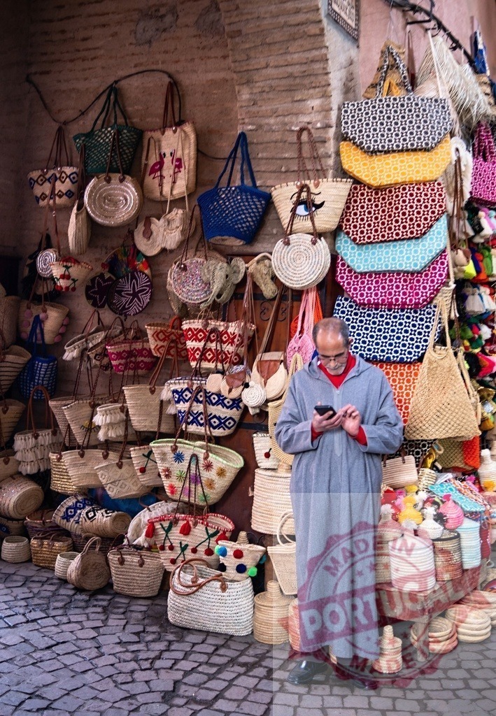 Moroccan shopkeeper using phone in front of his colorful handmade wicker bags shop | Marrakesh shopkeeper wearing traditional djellaba using mobile phone in his colorful wicker bag store in a traditional souk - Realized with Pictrs.com