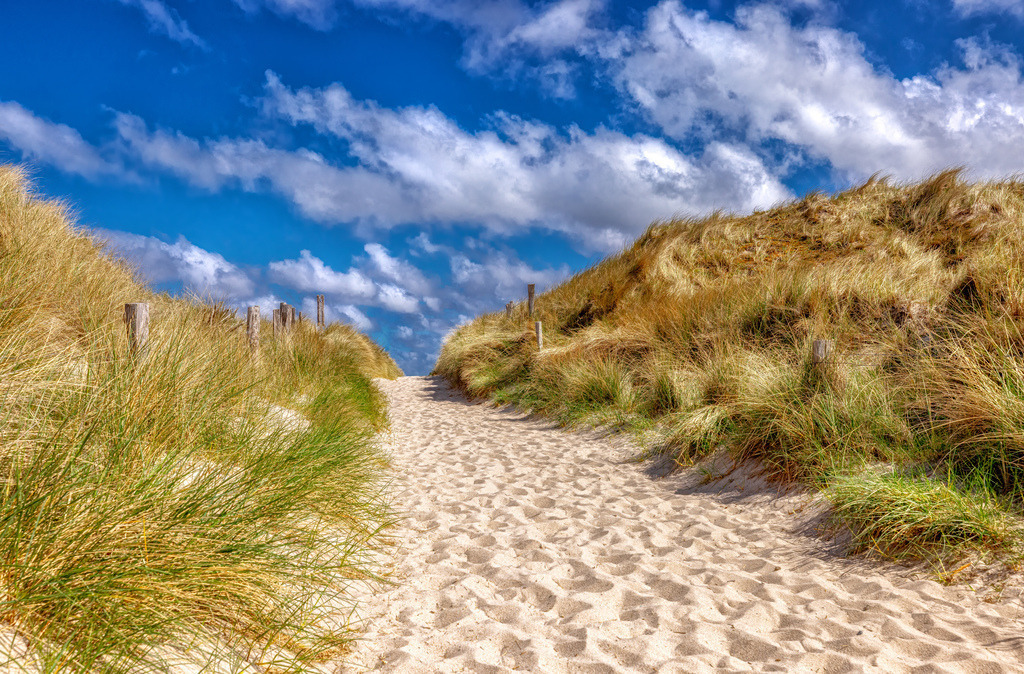 Malerischer Dünenweg durch Strandhafer-feinem Sand--Sylt I | Das Bild zeigt einen sandigen Dünenweg mit Strandhafer, der zum Strand führt.Die Szenerie ähnelt typischen Strandaufgängen an der deutschen Nord- oder Ostseeküste, wie etwa auf Sylt oder in Sankt Peter-Ording.Die Aufnahme zeigt eine sonnige Wetterlage mit blauem Himmel und weißen Wolken über der Dünenlandschaft. - Realisiert mit Pictrs.com