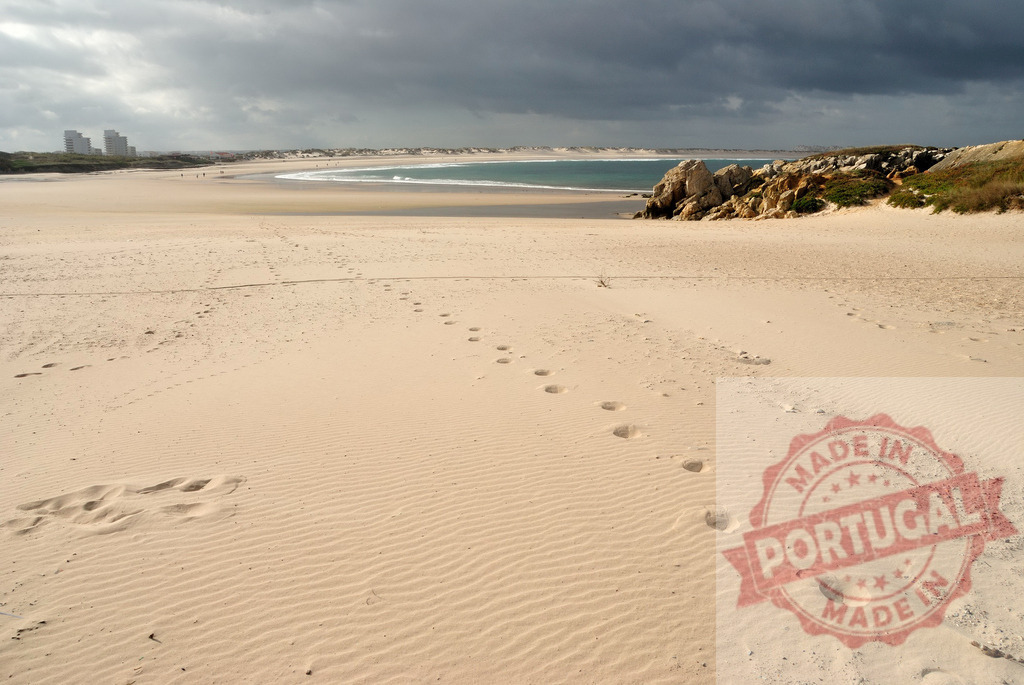 Footprints walking on sandy beach under storm clouds. Centro - Portugal | Footprints tracing a path on a vast beach leading towards the ocean and distant buildings - Realized with Pictrs.com