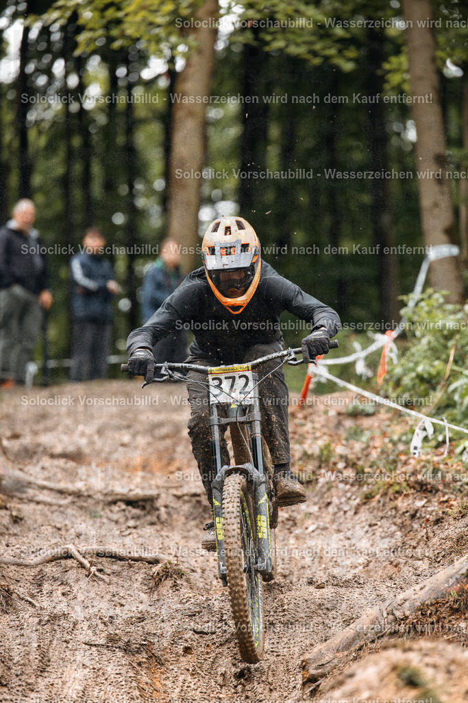 Mountainbike-Action im Wald | Ein Mountainbiker in voller Ausrüstung rast auf einem schlammigen, kurvigen Weg durch einen dichten Wald. Er trägt einen orangefarbenen Helm und eine dunkle Schutzkleidung. Der Fokus liegt auf dem Fahrer und seinem Mountainbike, während der Hintergrund verschwommen ist. Es scheint sich um einen Wettkampf oder Training in einem Gelände zu handeln. - Realisiert mit Pictrs.com
